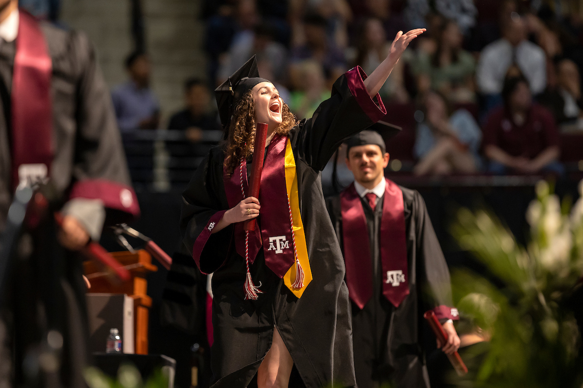 Texas A&M student in graduation cap and gown waiving to the crowd at a graduation ceremony.