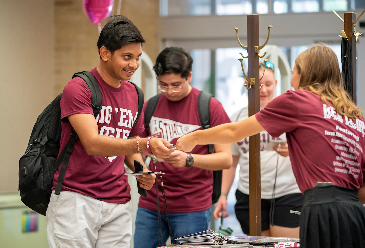 Students and visitors gather in a modern university library during an Open House event featuring informative displays and Aggie Spirit banners.